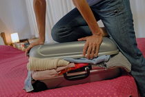 Woman trying to close overflowed suitcase on bed by Sami Sarkis Photography