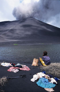 Girl washing clothes in a lake with the Mount Yasur volcano emitting smoke in the background von Sami Sarkis Photography